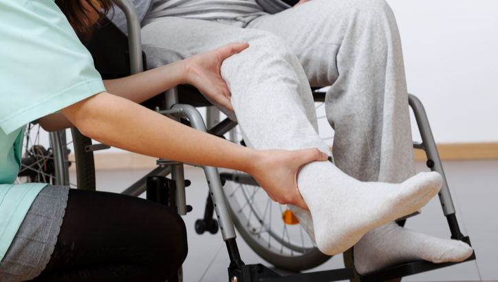 A healthcare worker supports and examines the leg of a person seated in a wheelchair.