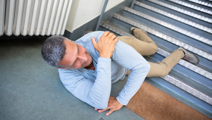A man lies on the floor at the bottom of a staircase, holding his shoulder and appearing to be in pain, suggesting he has fallen down the stairs.