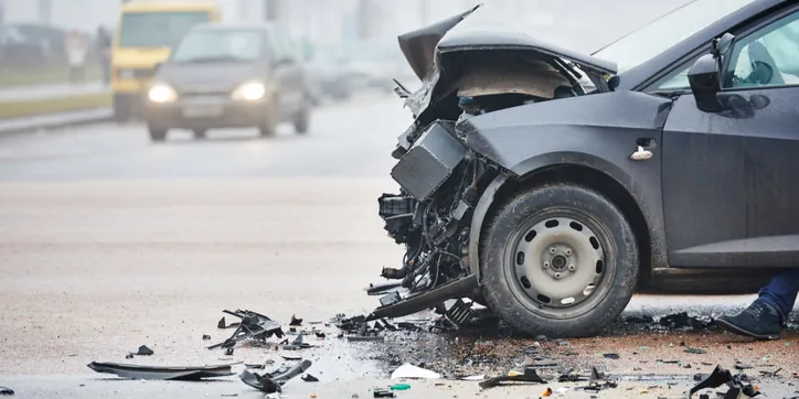 A dark-colored car with severe front-end damage is stopped on a road after a collision, with debris scattered around and other vehicles in the background.