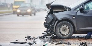 A car with severe front-end damage after a collision sits on a road, with debris scattered around and other vehicles in the background.
