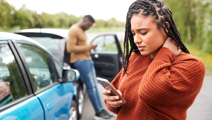 A woman holding her neck looks at her phone beside a blue car, while a man stands by an open car door using his phone.