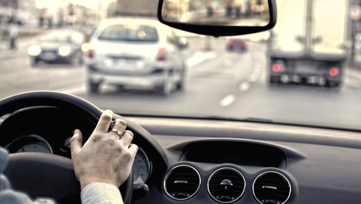 View from inside a car showing a driver's hand on the steering wheel while driving on a busy multi-lane road with several vehicles ahead.