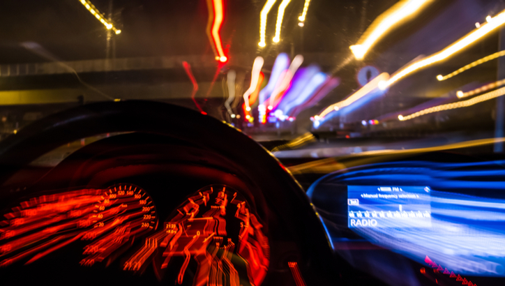 Blurred, colorful light trails seen from behind a car dashboard and steering wheel at night, suggesting motion or speed.