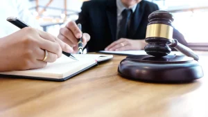 Two people sit at a desk with a gavel, one writing in a notebook and the other pointing, suggesting a legal or consultation meeting.