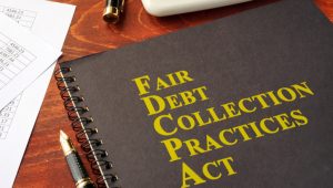 A black spiral notebook labeled "Fair Debt Collection Practices Act" in yellow text on a wooden desk with papers, a pen, and a gold item nearby.