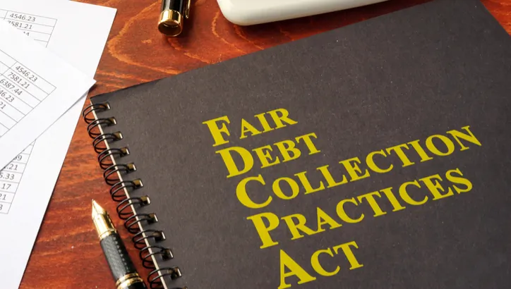 A black spiral notebook labeled "Fair Debt Collection Practices Act" rests on a desk with a pen, papers, and a brass object nearby.