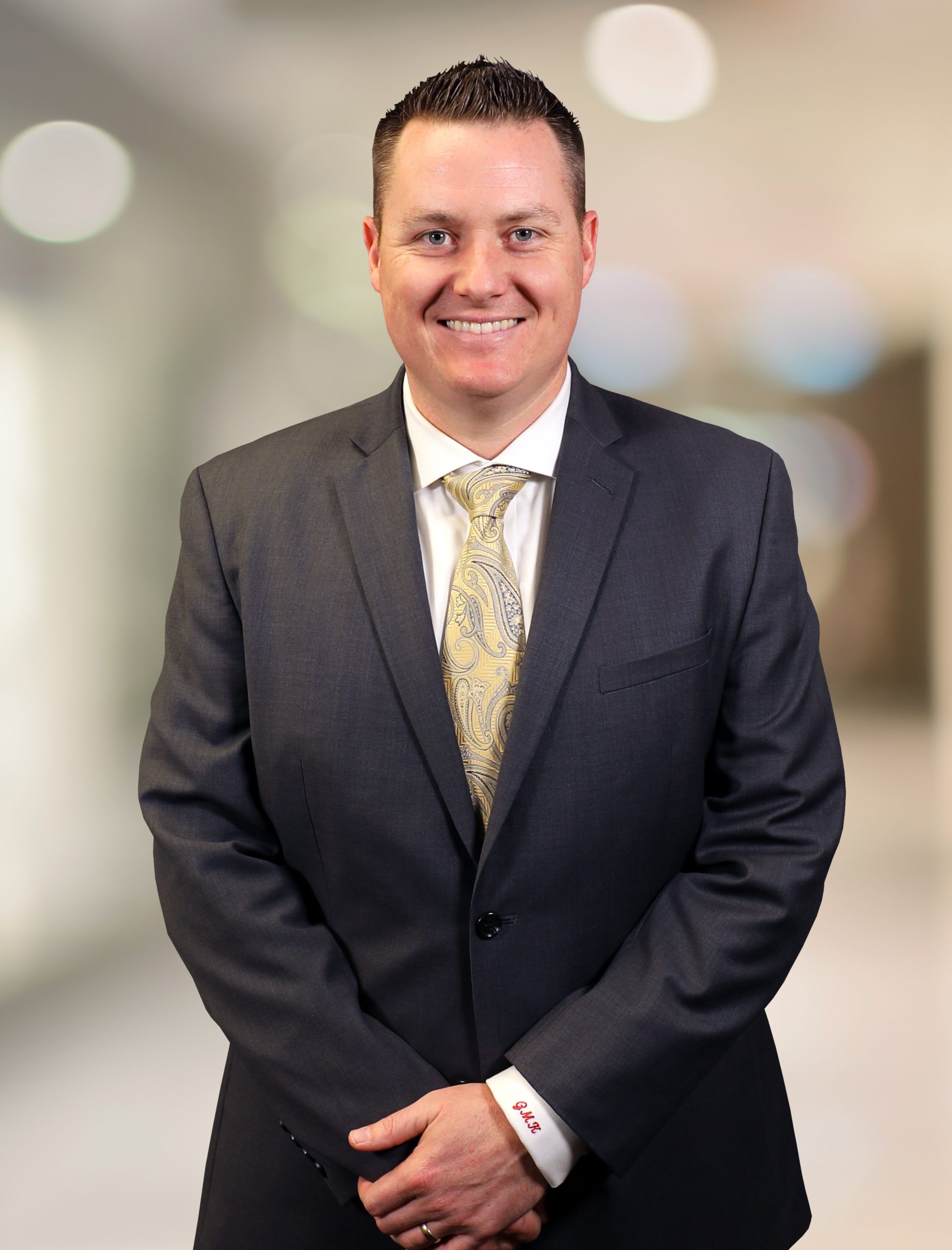 A man in a dark suit and paisley tie stands smiling in front of a blurred indoor background, with his hands clasped in front.