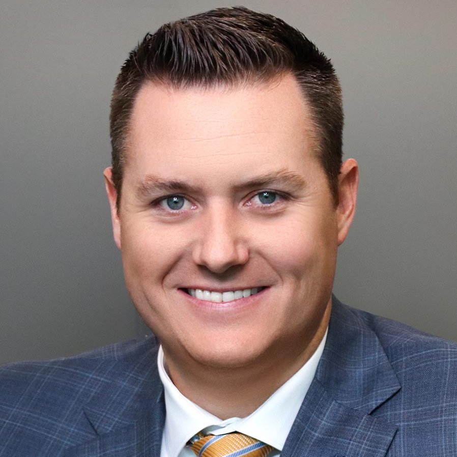 Smiling man in a blue plaid suit jacket, white shirt, and yellow striped tie, posing in front of a plain gray background.