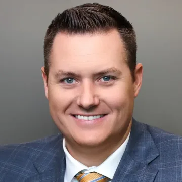 Smiling man in a blue plaid suit jacket, white shirt, and yellow striped tie, posing in front of a plain gray background.