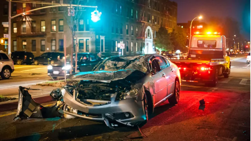 A heavily damaged silver car sits on a city street at night as a tow truck prepares to remove it. Traffic and apartment buildings are visible in the background.