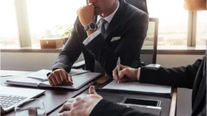 Two people in business suits sit at a desk with notebooks, one writing and the other with a hand on his chin, in an office with large windows.