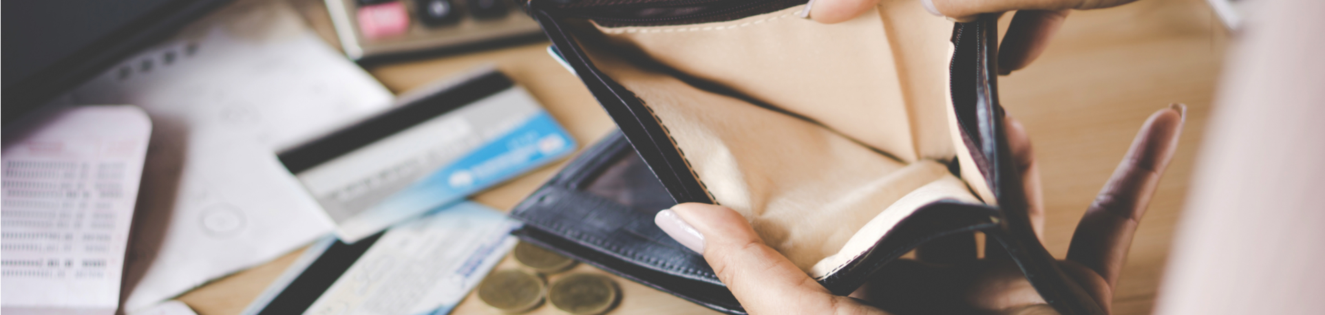 A person holds open an empty wallet on a table with credit cards, coins, and papers scattered nearby.