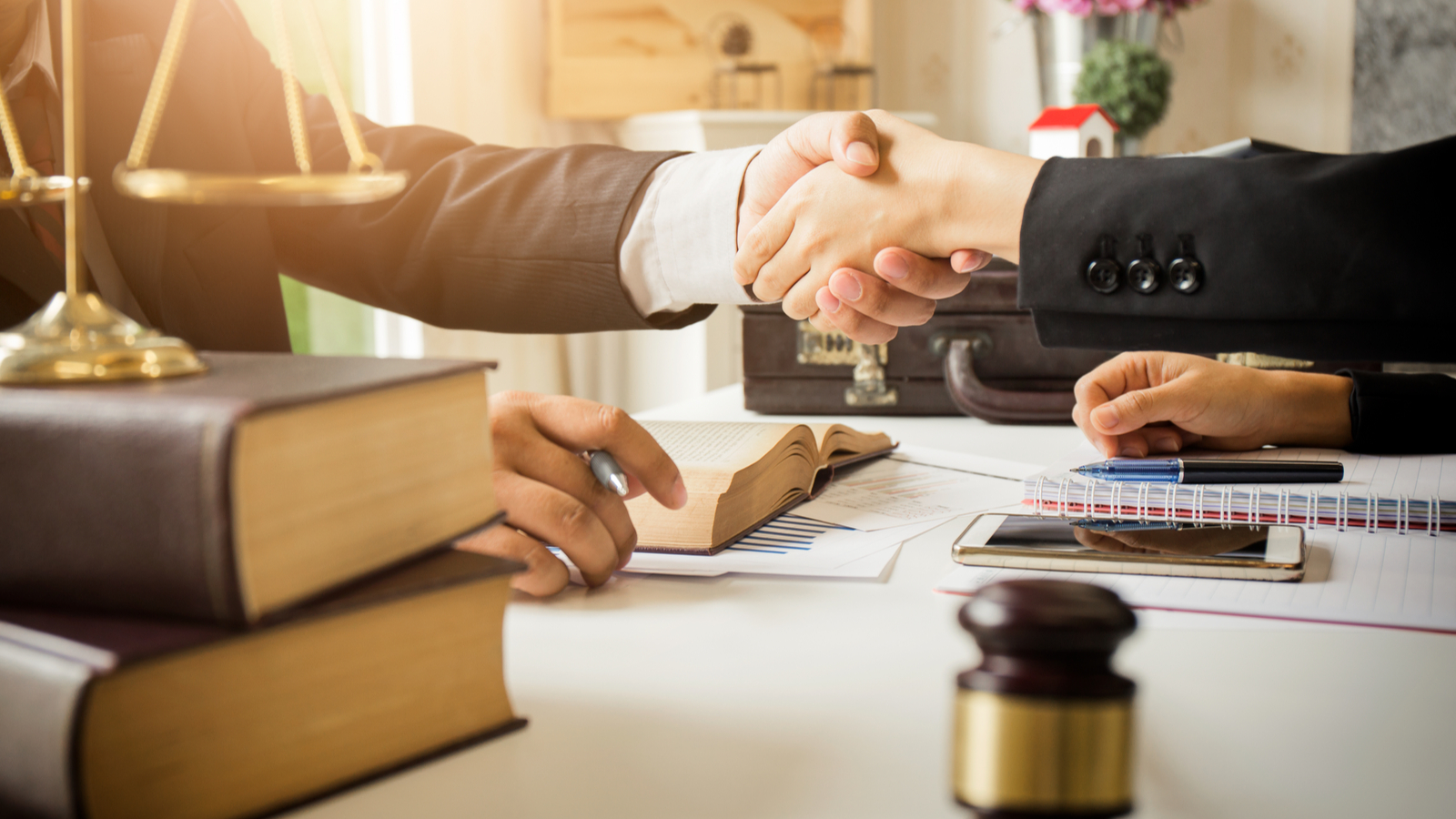 Two people in business attire shaking hands across a desk with legal books, documents, a pen, a gavel, and a smartphone visible.