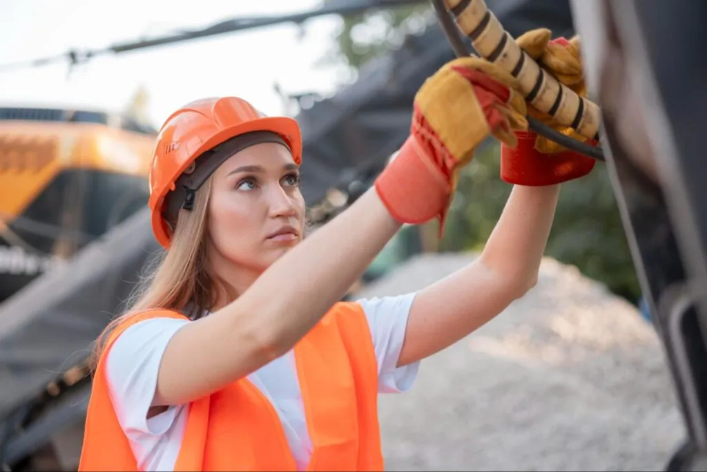 A woman in a hard hat, safety vest, and gloves works on machinery at a construction site.