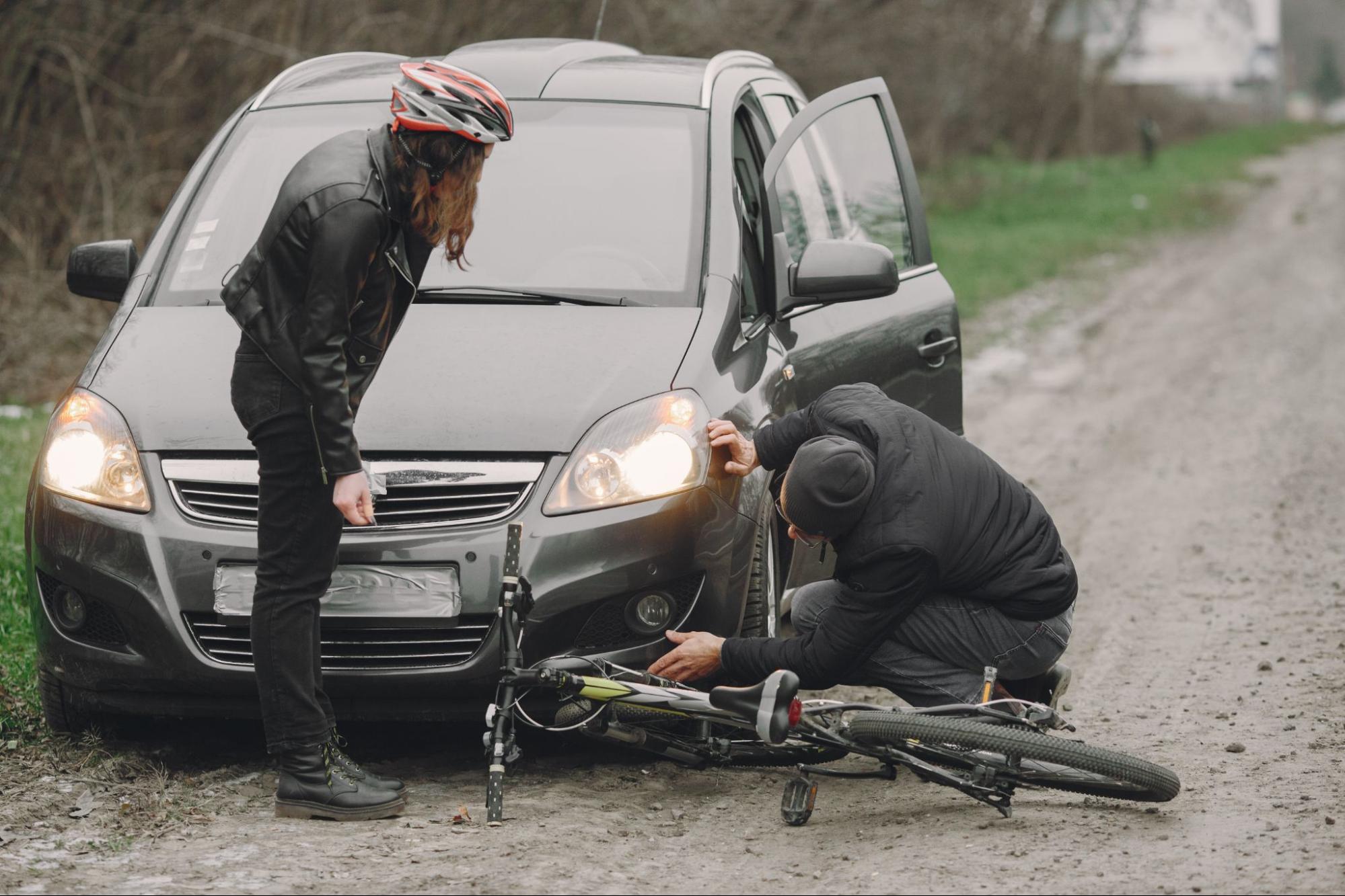 A person in a helmet stands by a car while another inspects a damaged bicycle lying on the ground in front of the vehicle on a dirt road.