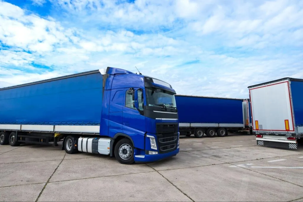A blue semi-truck with a trailer is parked on a paved lot beside other trailers under a partly cloudy sky.