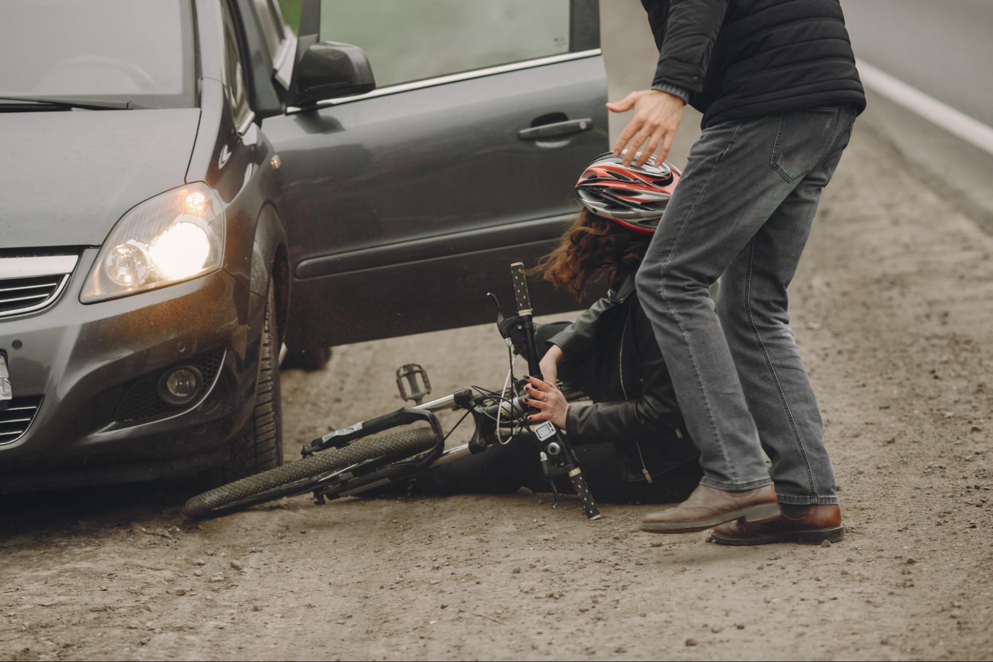 A person wearing a helmet sits on the ground next to a fallen bicycle and an open car door, while another person stands nearby.