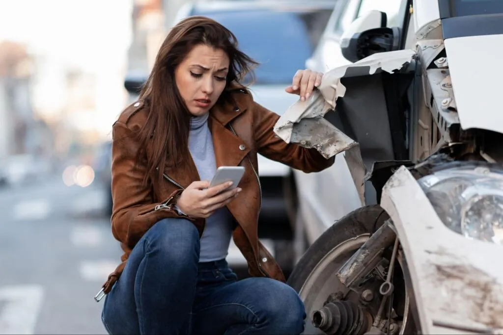 A woman crouches beside a damaged car, looking concerned while holding her phone, after a vehicle accident on a city street.