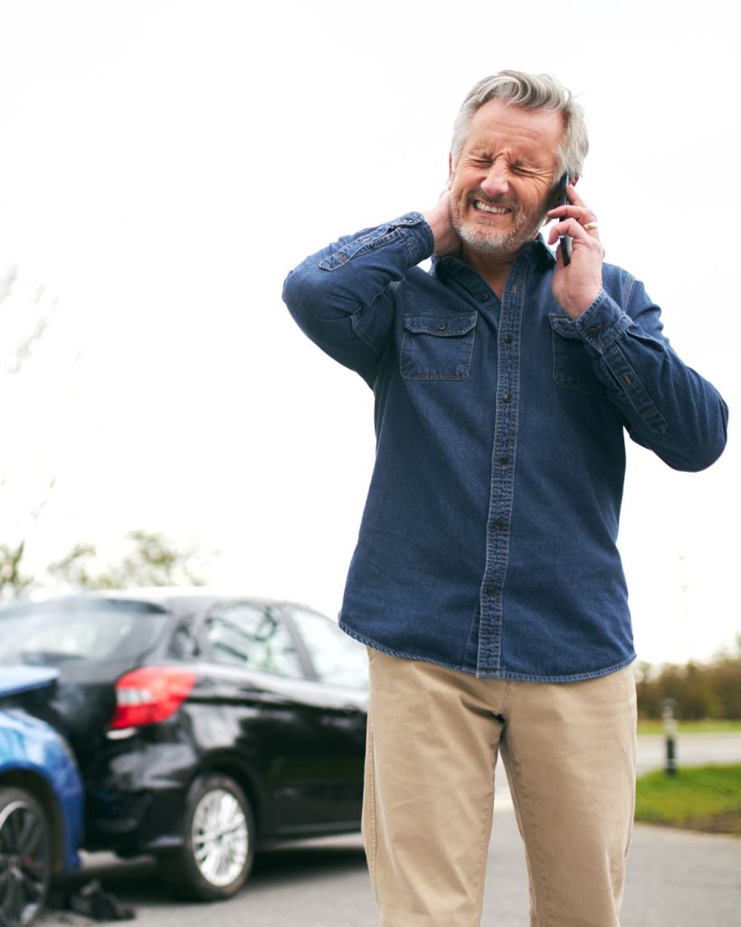 A man stands next to two damaged cars, holding his neck in pain and speaking on the phone, likely after a car accident.
