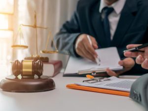 Two people in suits sit at a desk with legal documents, a gavel, scales of justice, and a law book, suggesting a legal or consultation setting.