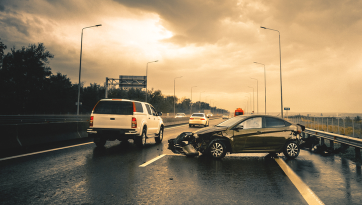 Black car with front-end damage positioned at an angle on wet highway; other vehicles pass by under cloudy, overcast sky.