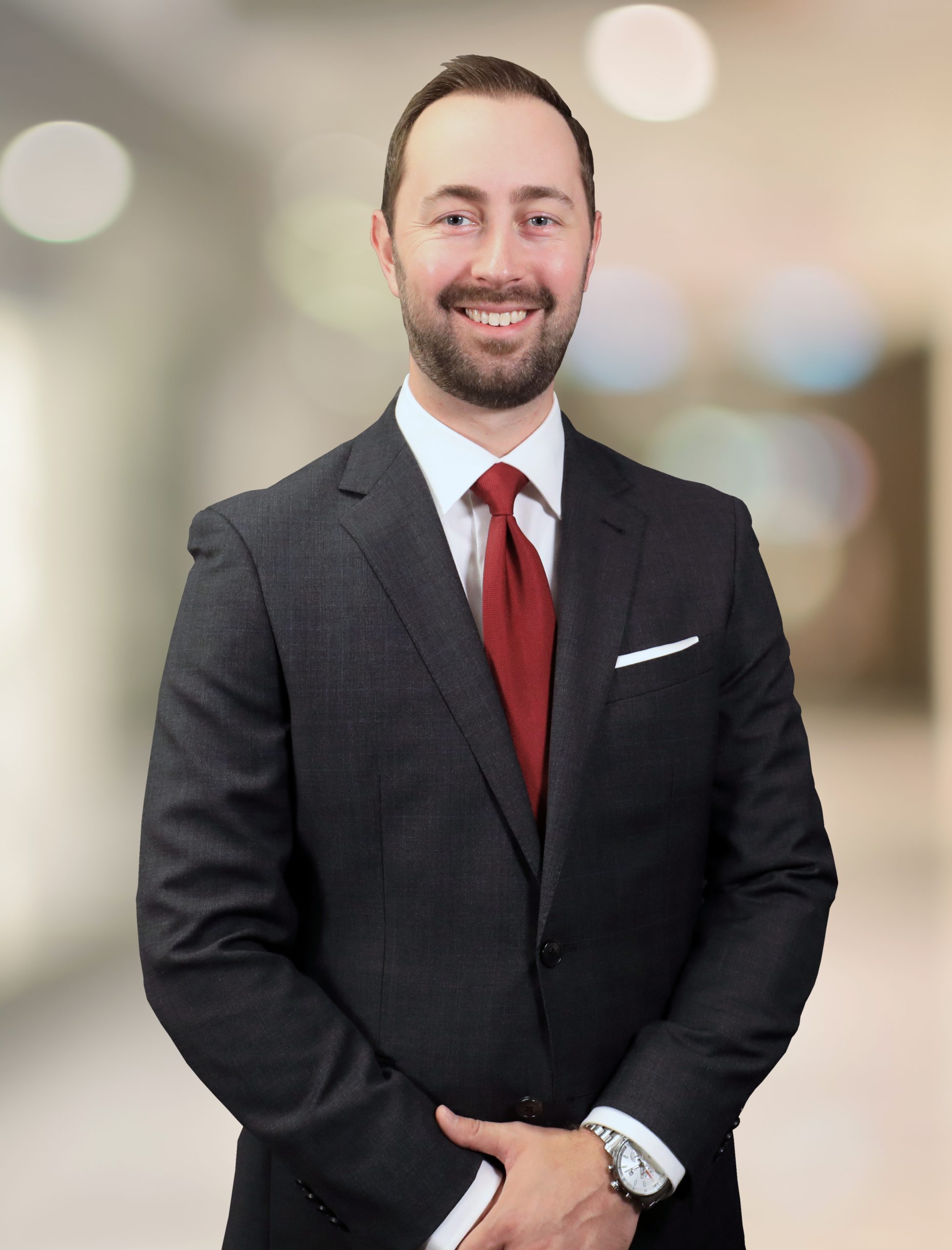 A man in a dark suit with a red tie and white pocket square stands smiling in front of a blurred indoor background.