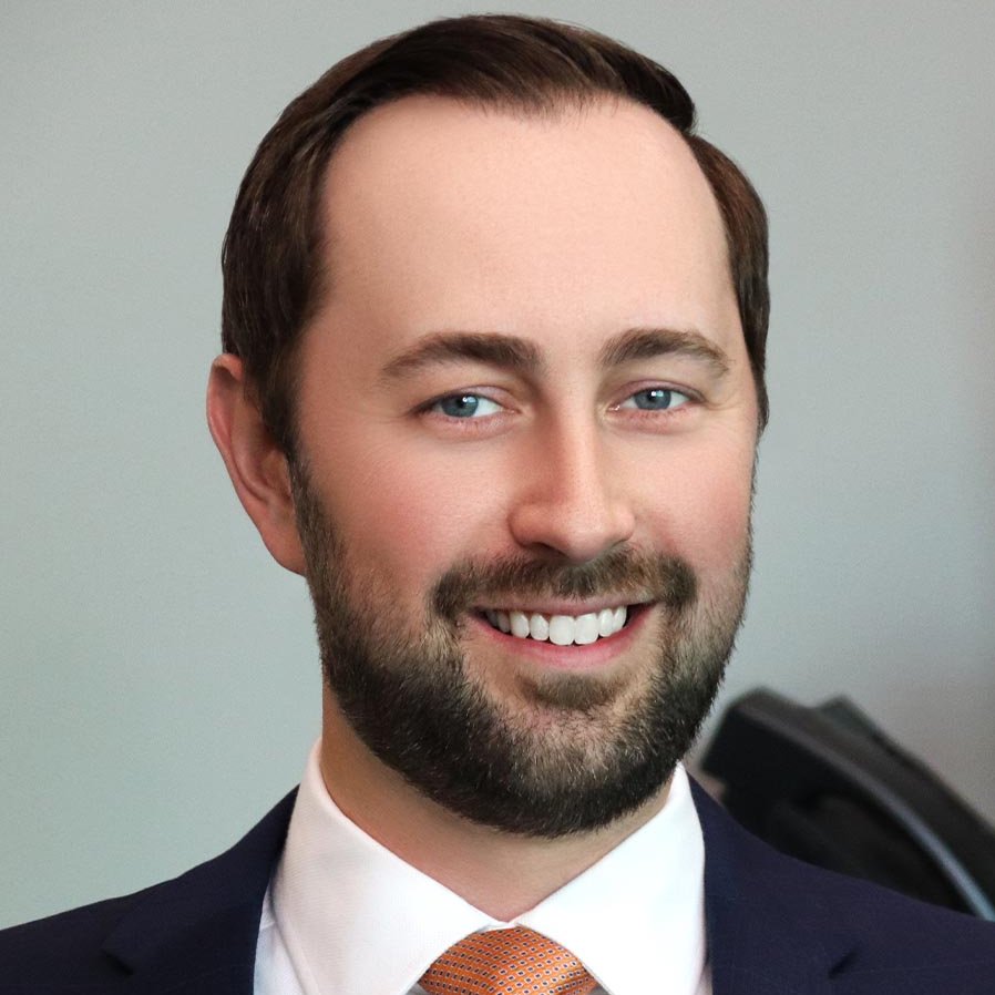 A man with short brown hair and a beard, wearing a dark suit, white shirt, and orange tie, smiling in front of a plain background.