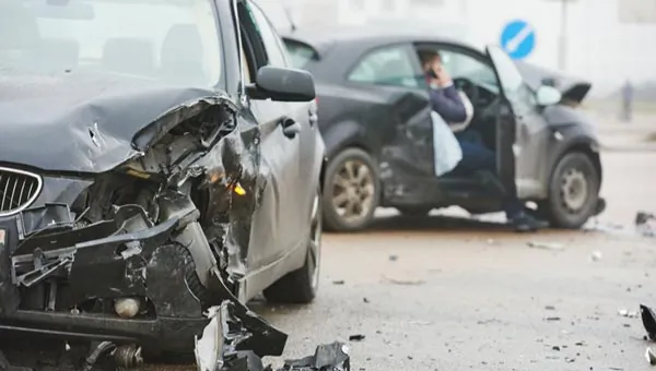 Two damaged cars are shown after a collision, with debris on the road and a person sitting inside one of the cars, talking on a phone.