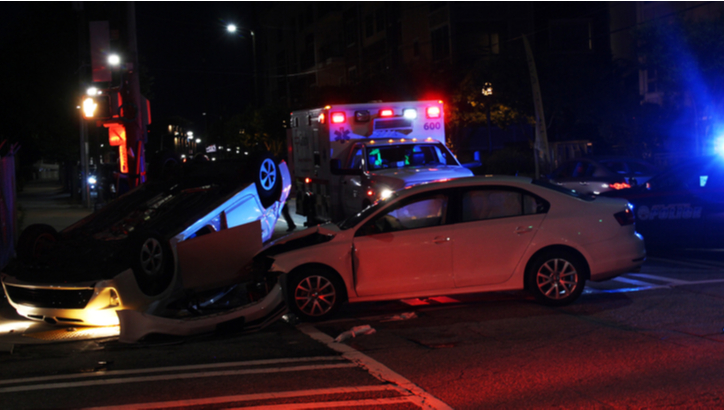 A nighttime scene showing a car accident at an intersection, with one car overturned and another damaged; police and ambulance vehicles are present with lights flashing.