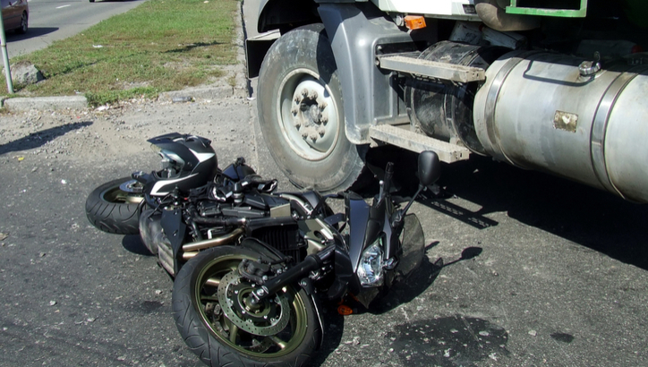 A motorcycle lies on its side beneath the front section of a large truck after a collision on a paved road.