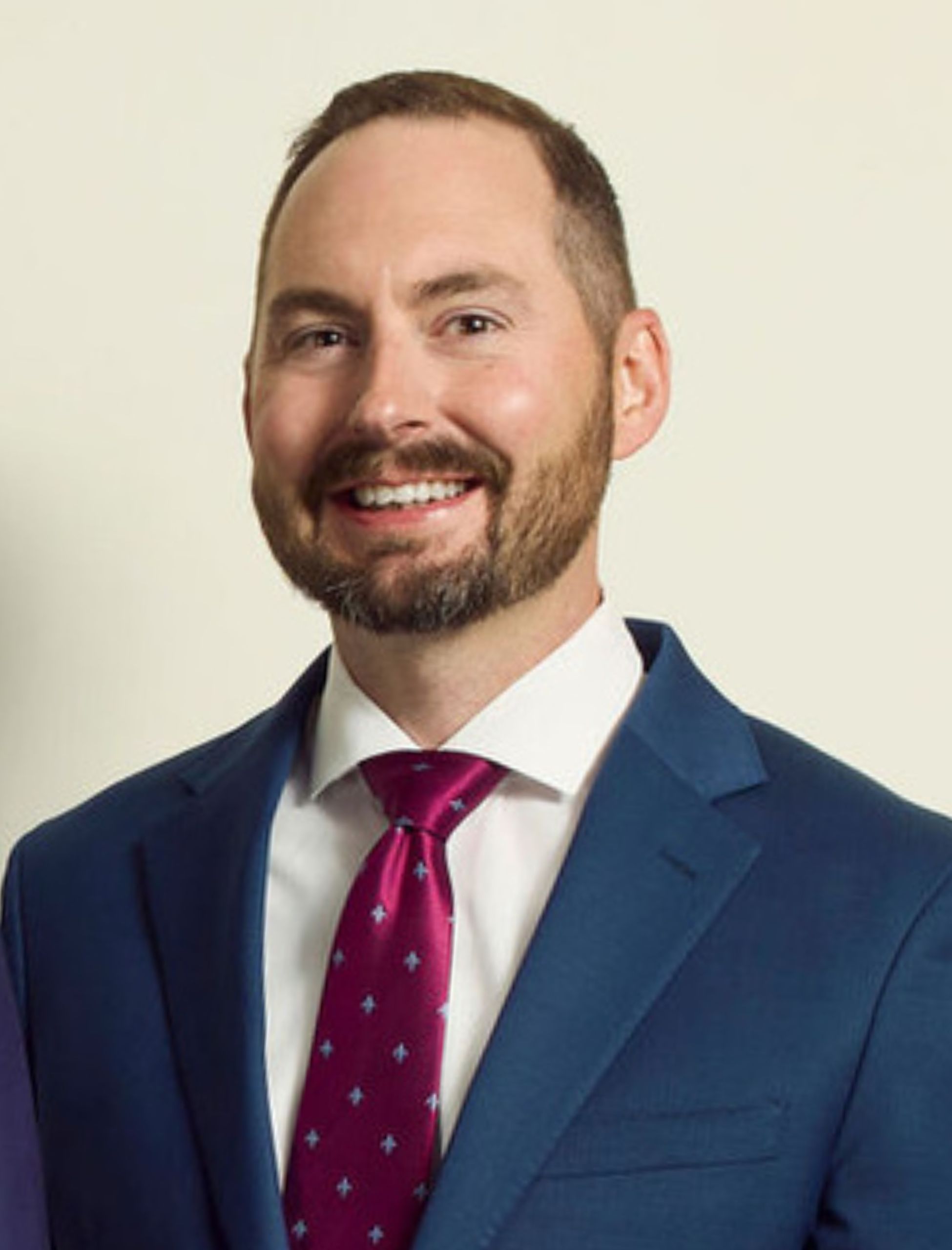 A man in a blue suit, white shirt, and red patterned tie smiles in front of a plain light background.