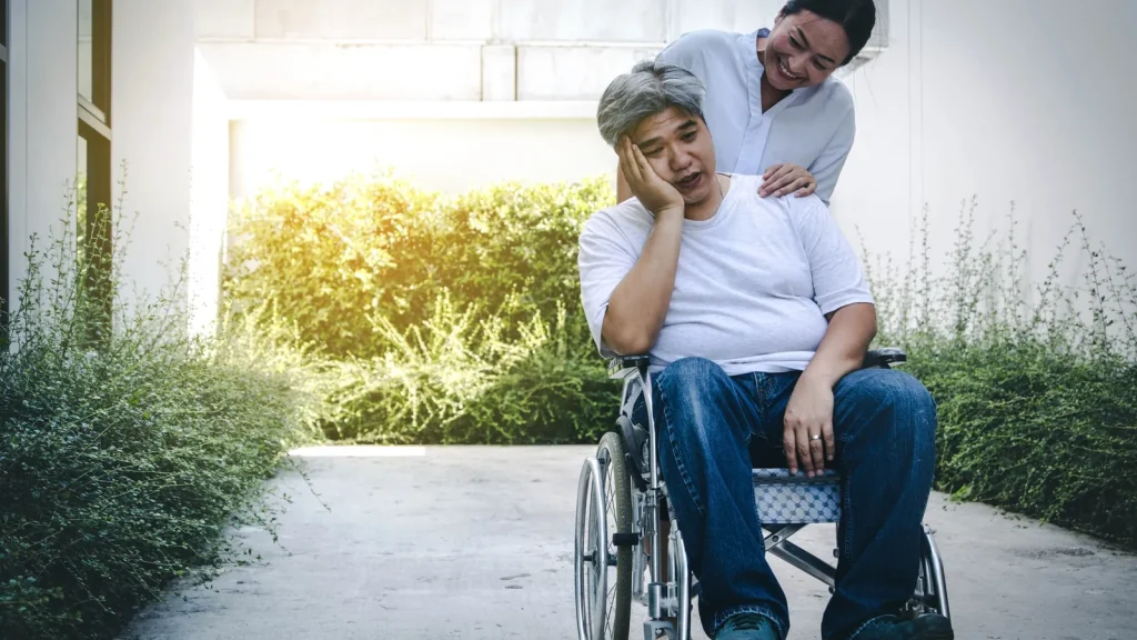 An older man sits in a wheelchair looking tired while a younger person stands behind him, smiling and placing hands on his shoulders in an outdoor setting.