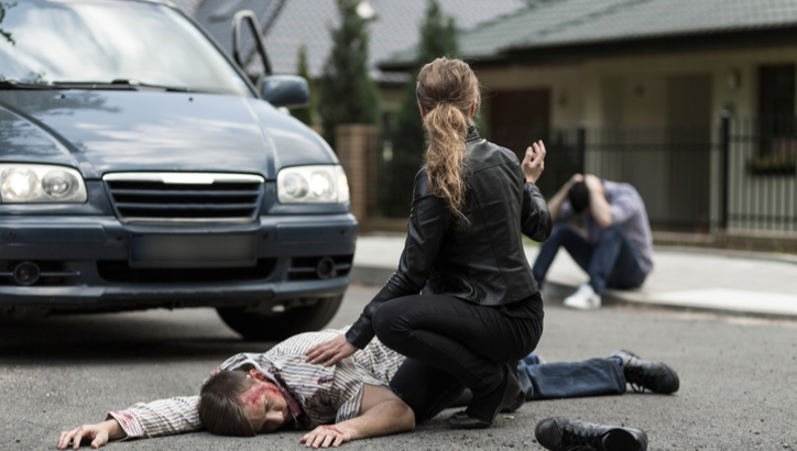 A woman kneels beside an injured man lying on the road in front of a car, while another person sits in the background holding their head.