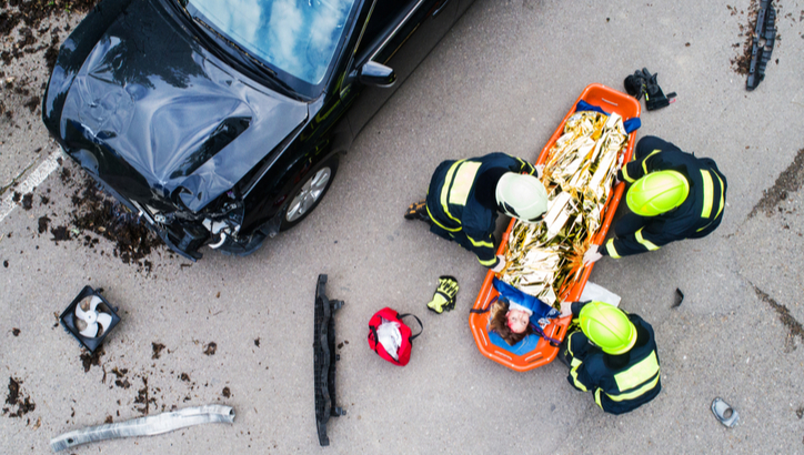 Three emergency responders assist an injured person on a stretcher next to a damaged black car at an accident scene.