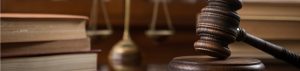 A wooden gavel rests on a sound block, with stacked legal books and a balance scale visible in the background.