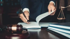 A person in a suit writes in a book at a desk with legal scales, a gavel, and stacked documents, suggesting a legal or law-related setting.