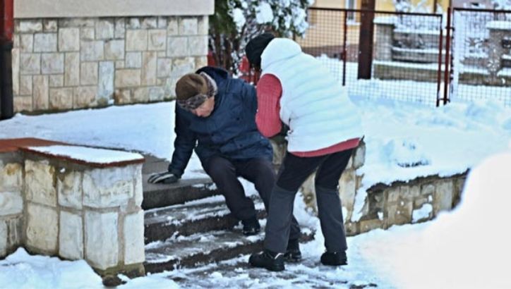 Two people are outside on snowy steps; one person is sitting on the steps while the other helps them up. Snow covers the ground and surrounding area.