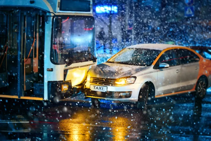 A taxi and a city bus collide on a snowy street at night, with snow falling heavily and headlights illuminating the scene.