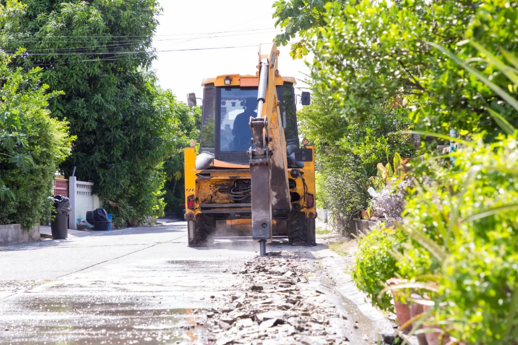 A yellow backhoe excavator breaks up pavement on a residential street, with debris scattered and greenery on both sides.