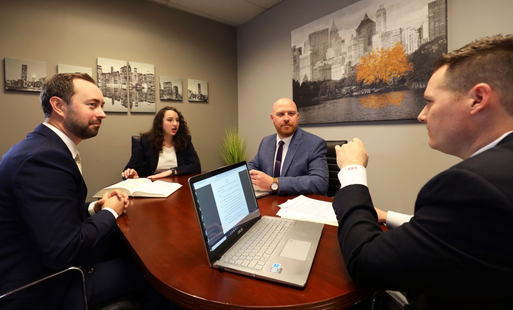 Four people in business attire sit around a conference table with documents and a laptop, engaged in a discussion in an office with cityscape artwork on the wall.