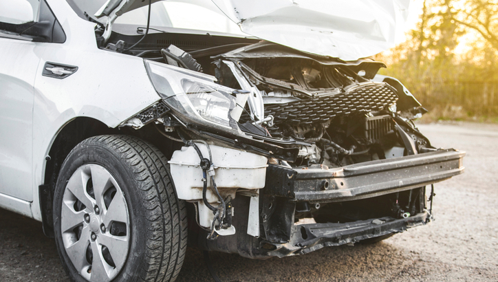 A white car with severe front-end damage, including a crumpled hood and exposed internal parts, parked on a paved surface outdoors.