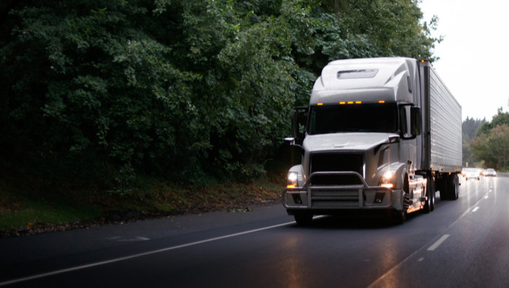 A large silver semi-truck drives on a wet road bordered by dense green trees, with several vehicles visible in the background.