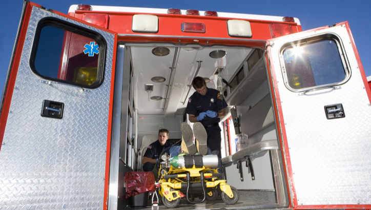 Two paramedics are inside an ambulance with open rear doors, preparing a stretcher with medical equipment visible inside the vehicle.