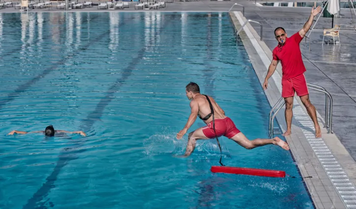 A lifeguard jumps into a swimming pool with a rescue tube as another lifeguard signals, while a person floats face down in the water.