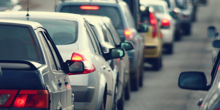 A line of cars is seen in traffic on a road, with brake lights illuminated and vehicles closely packed together.
