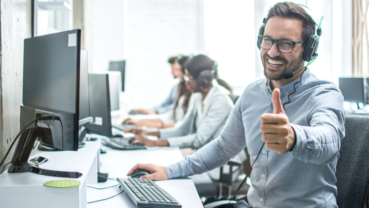 A man wearing a headset sits at a desk with a computer, smiling and giving a thumbs-up. Other headset-wearing coworkers are seated in the background.