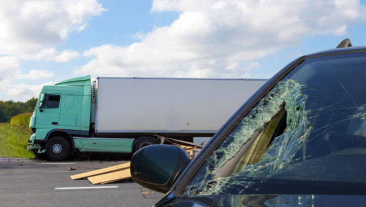 A damaged car with a shattered windshield sits near a large truck that has veered off the road, with debris scattered between them.