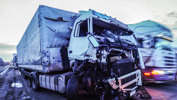 A large truck with severe front-end damage is parked on the side of a road, while another truck passes by in the background.