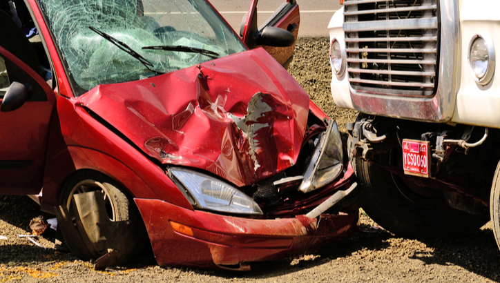 A red car with a crumpled front end is crashed into the front of a large white truck on a gravel road.