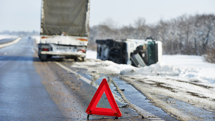 A red warning triangle is placed on a snowy road near an overturned truck, with another truck parked ahead.